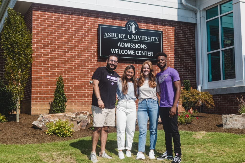 Four students in front of Corbitt Hall at Asbury University, the brick with white trim Admissions Welcome Center.