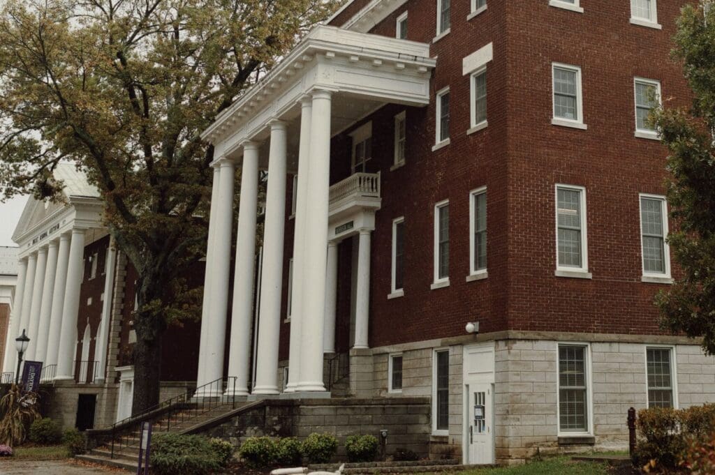 Side exterior of Morrison Hall at Asbury University with maroon brick, white trim, flat roof, and front portico with columns.
