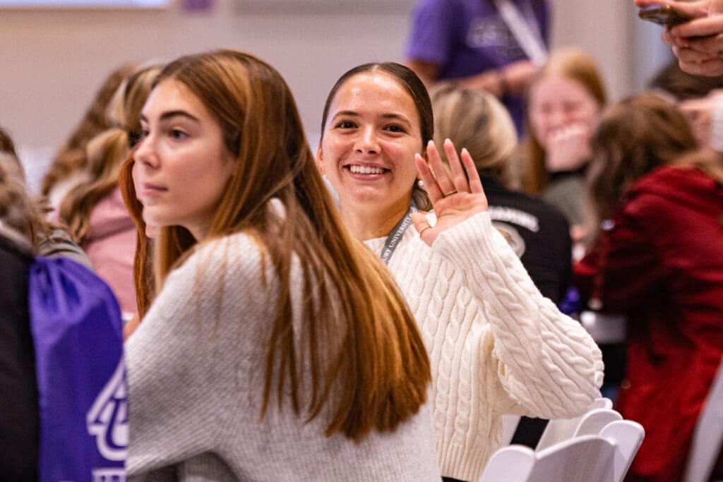 Two girls stting at table. One is looking at camera smiling and waving. Event is Asbury University's Educator's Rising Regional Experience.