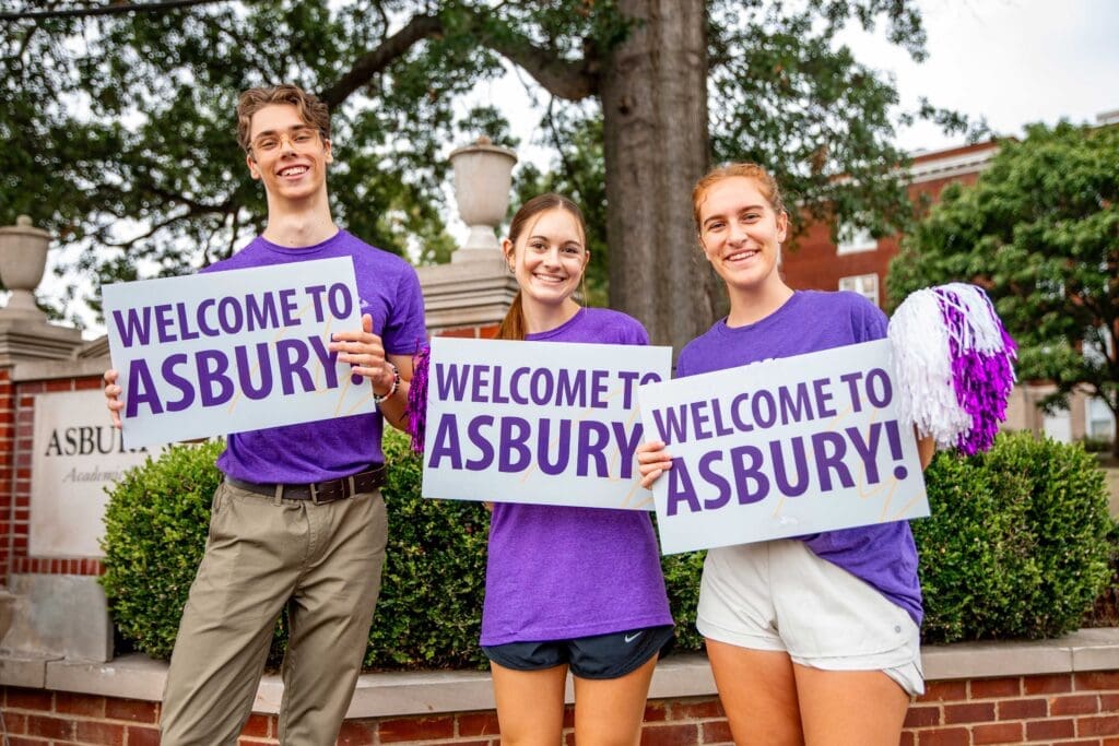 Three students wearing Asbury apparel and holding welcome signs for freshmen move in day Asbury University.