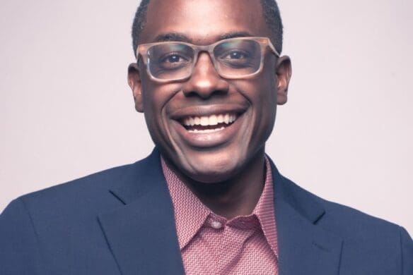 headshot of Shawn Okpebholo, smiling in a suit jacket wearing glasses