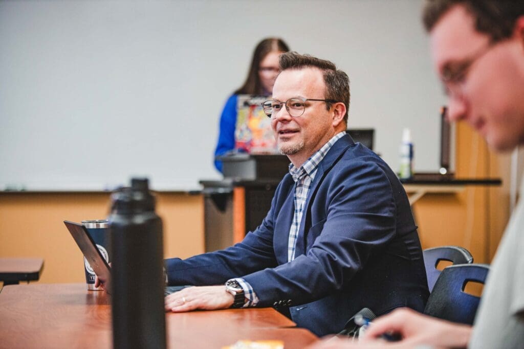 Man in business suit sits at a table with other students