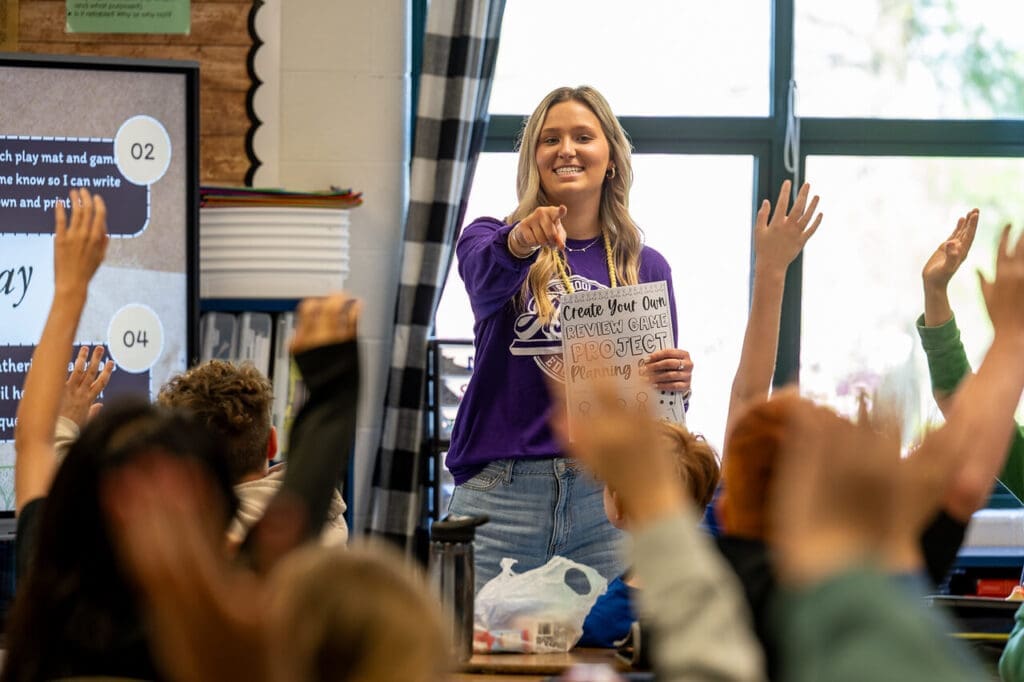 Asbury education student calling on a student with a smile in a classroom of kids with their hands raised.