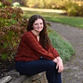 Female Asbury media communications student Emily Hurt sits outside on a low wall in a red top and jeans