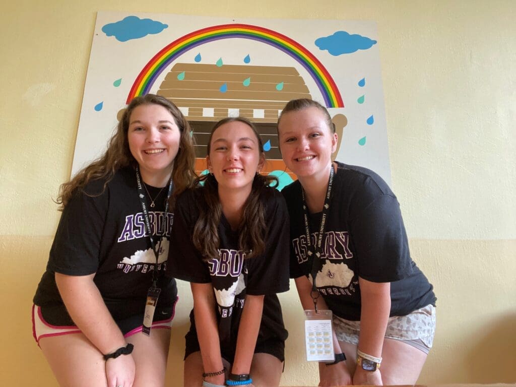 Three female AU students on a mission trip smile in front of classroom artwork